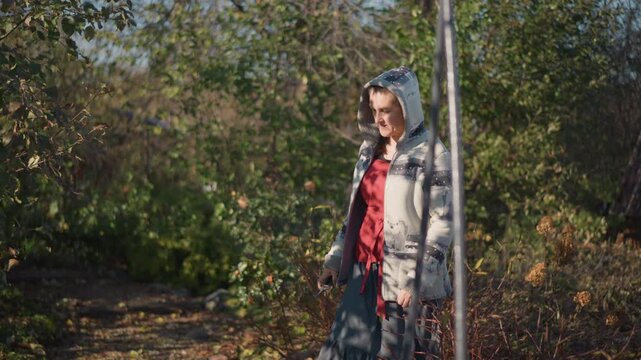 Garden gate white woman harvesting produce. Hooded gardener pauses by rustic fence and shed, carrying basket and tools, picking vegetables and fruit near cottage, tactile soil and branch details, slow
