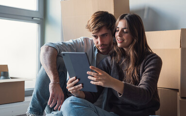 A young couple sits among cardboard boxes in their new apartment, smiling as they use a tablet to...