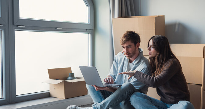 Young couple sits on the floor of their new apartment surrounded by cardboard boxes, using a laptop to plan moving tasks, sort finances and discuss decorating and unpacking together