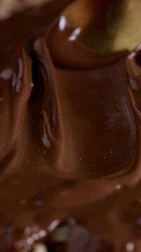 Close-up of melted chocolate being spread over hazelnuts with a spoon. Smooth glossy texture and dessert preparation process.