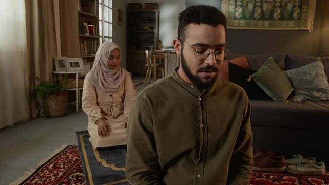 Medium shot of young Muslim couple praying together at home performing daily religious ritual with focus on bearded man finishing prayer in foreground, copy space