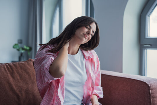 A young woman sits on a sofa massaging her neck and wincing from pain or stiffness. Casual clothing, soft natural light and a calm living room emphasize tiredness, stress and self-care