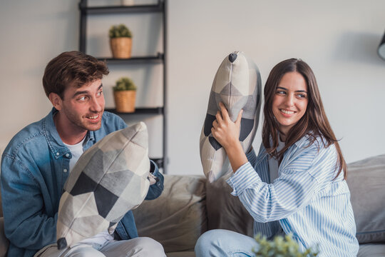 A smiling young couple enjoys a playful pillow fight on a cozy sofa in their living room, sharing laughter and relaxed moments that convey intimacy, fun and comfortable everyday life at home.