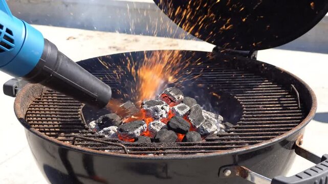 Igniting charcoal briquettes with a leaf blower creating sparks in outdoor cooking grill before grilling