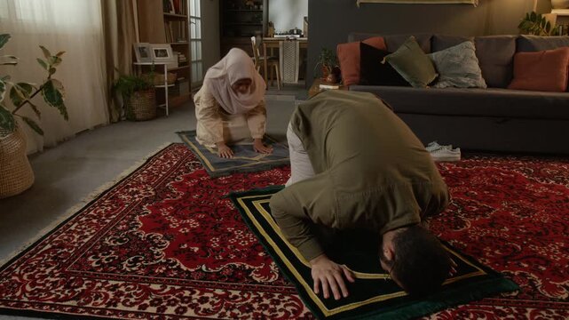 Full length shot of young Muslim couple praying and performing Salah in daily religious ritual sitting on prayer mats with focus on bearded man in foreground, copy space