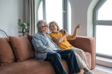 Obraz premium Warm senior couple relax on a sofa in their bright living room, smiling and sharing a joyful moment as the woman points toward a window. Comfortable, affectionate, and peaceful home scene.
