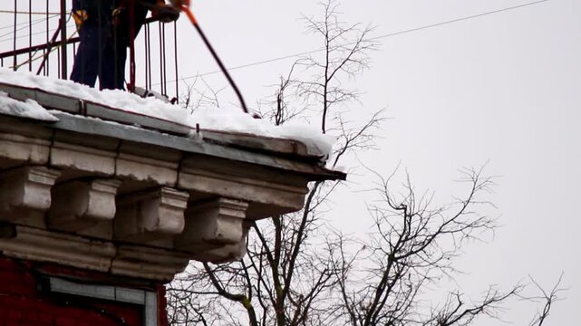 A worker removes snow from the roof of a building using a long-handled shovel