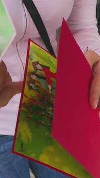 A woman shops for pop-up cards at a market in Hoi An, Vietnam. She is holding a lotus flower card and a bicycle card, admiring the craftsmanship and detail.