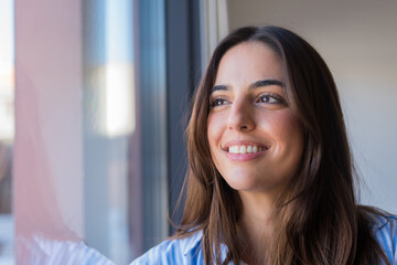 Obraz premium Smiling young woman looking out window with natural light, hopeful relaxed portrait in home