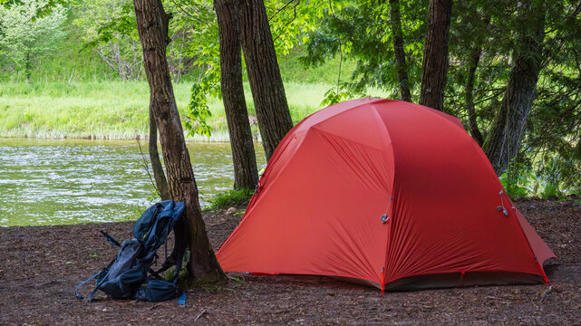 Eye-level profile view of bright orange tent set up next to a river with a backpacking pack leaning against a nearby tree
