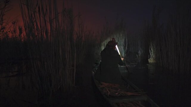 Lone poacher in a wooden boat searching for prey with a headlamp in a dark and eerie swamp