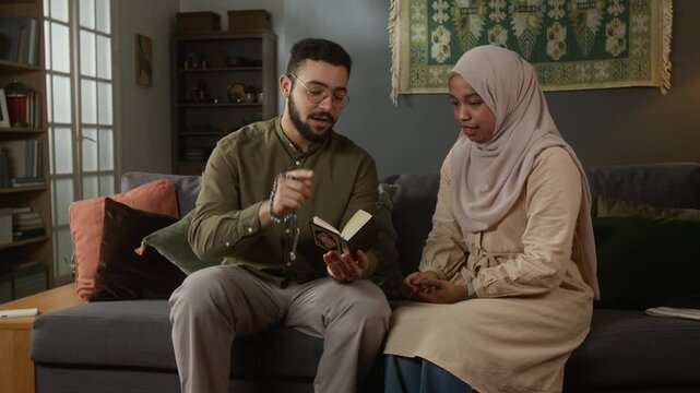 Front view shot of modern Muslim couple reading Quran together while dressed in modest clothing with man explaining verses to wife sitting on sofa at home