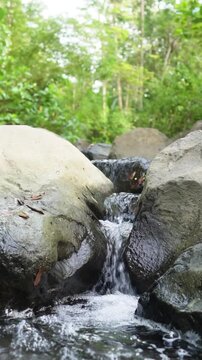 Tranquil Forest Waterfall Flowing Over Rocks With Soft Sunlight and Lush Greenery, peaceful nature vertical background. Flowing Mountain Stream Over Rocks Through Tropical Forest With Lush Green Folia