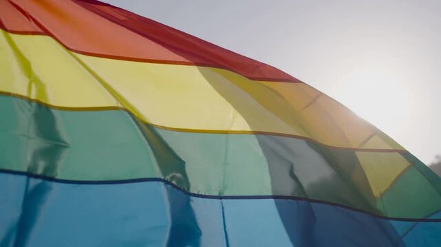 Fluttering rainbow flag rippling in light breeze over hills, with sun backlit fabric and flagpole