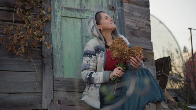 Caucasian homemaker sitting on rustic porch holding dried hydrangea, soft autumn light, weathered green door, knit coat and long skirt, serene reflective expression, quiet home moment