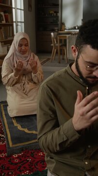 Vertical shot of Muslim young couple in daily prayer together sitting on prayer mats in modern home interior, focus flowing from man to woman