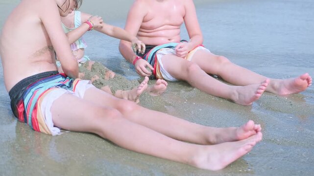 three children sibling sitting on sandy beach with feet in shallow ocean water, enjoying waves and summer fun together during family vacation, kids playing seaside with sand and water, childhood joy