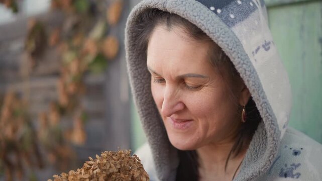 Caucasian woman holding dried hydrangea near green door, closes eyes to inhale scent, warm hoodie and soft grin, autumn leaves on weathered wood, homemaker and floral crafter enjoying seasonal craft