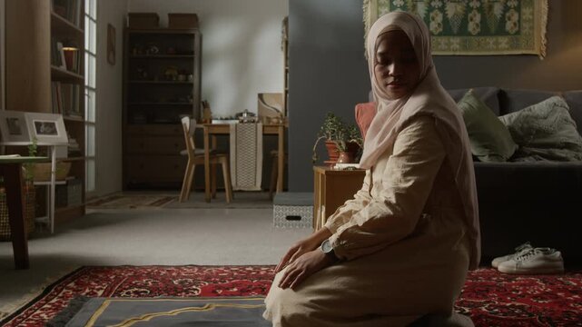 Side view shot of Muslim young woman finishing daily prayer ritual at home, turning head during salah in modern home interior, copy space