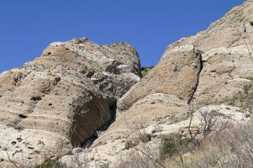 Fototapeta premium Large rugged tan rocks rise against a clear blue sky with sparse vegetation at their base creating a dramatic and dry mountainous landscape full of natural formations