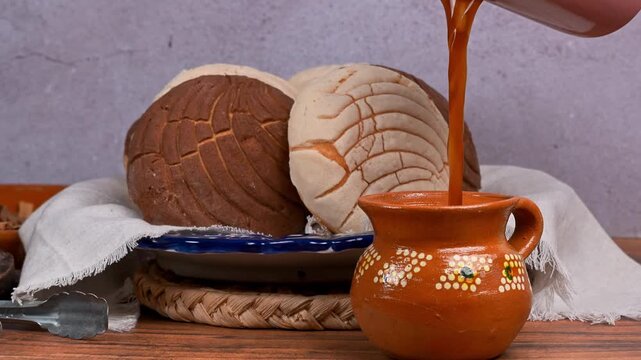 Serving coffee in a clay cup, with an assortment of Mexican bread, chocolate and vanilla conchas in the background