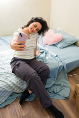Fototapeta premium Young woman smiling, sitting on a bed in her new first apartment and taking a selfie with a smartphone