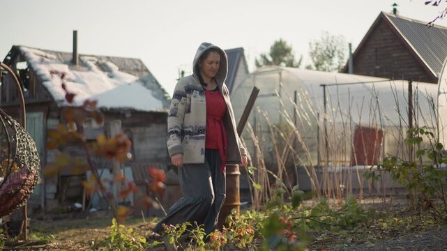 White woman walking in home garden, cozy autumn homestead scene with sweater and hoodie, strolling past rustic shed and barrel near vegetable beds, soft morning sunlight, calm reflective mood