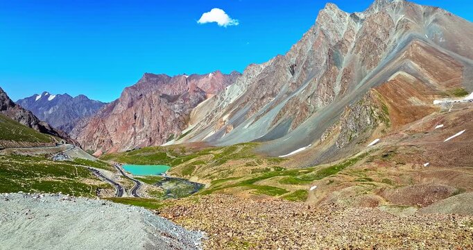 Dramatic aerial view of high mountain peaks and turquoise lake along a winding mountain pass road in Xinjiang, China.