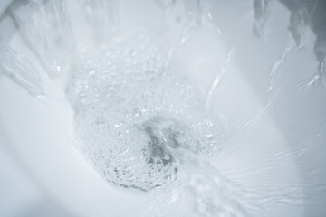 Water swirling in a white ceramic toilet bowl during a flush, foamy bubbles and motion blur forming a vortex around the drain, clean bathroom close up with bright highlights and minimal white interior