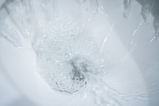 Water swirling in a white ceramic toilet bowl during a flush, foamy bubbles and motion blur forming a vortex around the drain, clean bathroom close up with bright highlights and minimal white interior