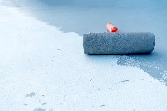 Close-up of a paint roller applying white liquid coating or sealant on a grey industrial floor. Construction site floor renovation and waterproofing process. Space for text on the surface.