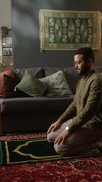Vertical side view shot of bearded Muslim man performing daily prayer on prayer mat and standing up after finishing ritual