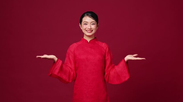 Smiling Asian woman gesturing both hands outward, red traditional outfit, friendly and welcoming mood on red background
