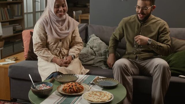 Zoom out shot of Muslim young woman maklouba dish on table and joining husband sitting on couch while enjoying traditional meal at home together