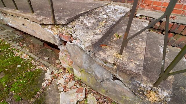 Crumbling concrete stair landing with a broken edge and scattered debris beside a brick wall and metal railing, showing building deterioration and a safety hazard.