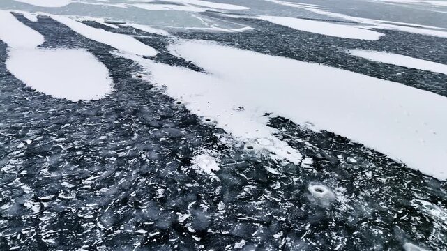 Aerial winter view of an ice fishing area with drilled holes across patterned frozen surface and snow patches, capturing cold weather recreation and risky thin-ice conditions.
