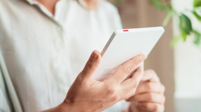Woman reading ebook on tablet device in bright room. Online learning, technology and comfortable home lifestyle concept with digital screen and green plants background