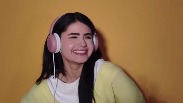 Cheerful beautiful woman with long dark hair wearing pink headphones, smiling and dancing while enjoying her favorite playlist against a vibrant yellow background in a studio setting
