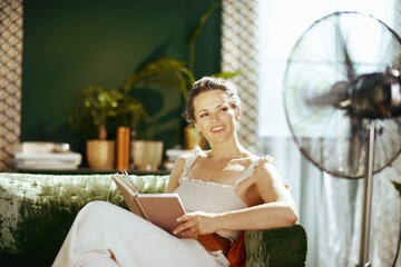 A middle-aged woman comfortably reads an open book on a green velvet sofa, smiling contentedly in her brightly lit living room with a standing fan. © Alliance