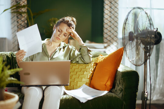 A mature woman on a green velvet couch looks exhausted, fanning herself with paper while working on a laptop. This scene captures the struggle of working from home amidst heat or stress.