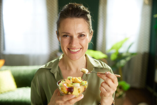 Happy woman holds a glass bowl of mixed fruit salad, savoring a spoonful of banana in her cozy, sunlit living room.