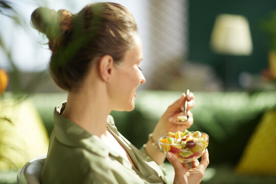 A woman in profile smiles while eating a mixed fruit salad in her living room. The cozy home interior and natural light highlight a moment of healthy snacking and self-care.