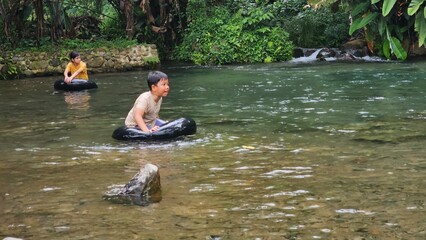 Fototapeta premium Two east asian kids ride a swim ring or swimming tyre and playing happily at a clean and shallow river. sstkTravel