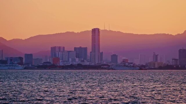 A timelapse view of the George Town urban skyline during sunset, featuring the prominent Komtar Tower and cruise ships sails across the sea.