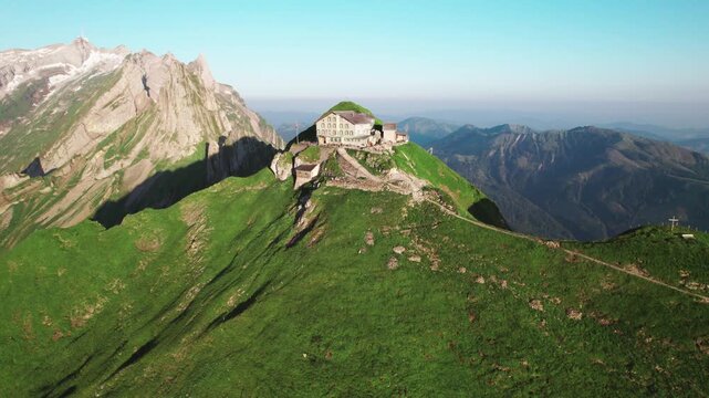 Aerial 4k drone view of the Schaeffler Hut perched on a lush green ridge, surrounded by rugged mountain peaks and sweeping alpine vistas under clear summer skies in Switzerland