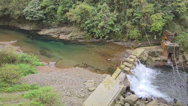 Small waterfall and concrete dam on a clear forest river with lush green foliage.