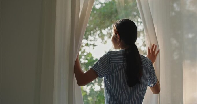 Asian woman standing by the window and happily opening both curtains to let the soft natural morning light into her house. 