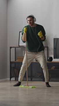 Young man happily dancing with mop while listening to music on headphones during fun house cleaning session in modern living room interior with wooden floor