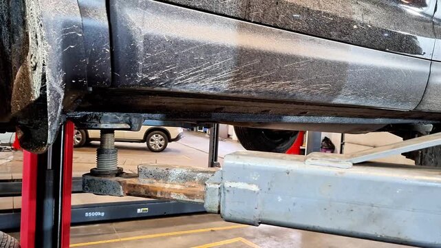 Underside view of vehicle elevated on hydraulic lift in automotive repair shop, showing lift arms supporting frame with background equipment, highlighting maintenance and inspection process