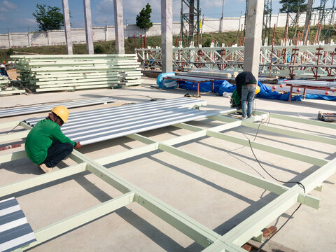 workers assembling and securing steel structure frame, attaching metal siding sheets to form walls of industrial building. Industrial construction, warehouse development and factory building projects.